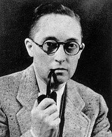 A black and white picture of Alan Hart smoking a pipe. He wears a light coloured suit and has on circular glasses.